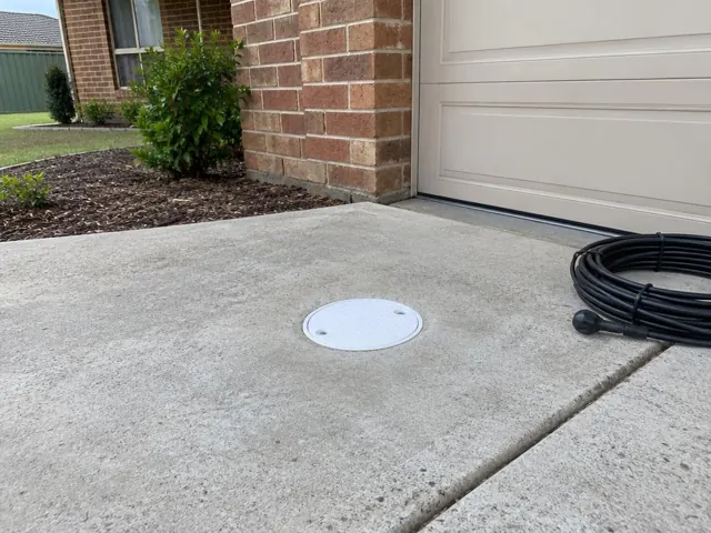 Capped drain inspection opening on a concrete driveway beside a coiled camera reel, brick home facade in Mount Druitt