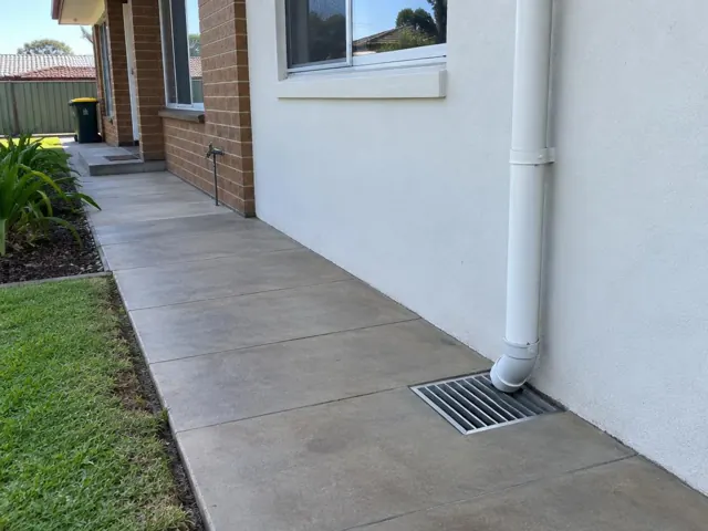 Outdoor gully grate beside a downpipe on a paved side path and brick house in Plumpton